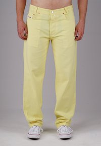 Yellow denim pants with a relaxed fit, featuring five pockets, a zip fly, and metal hardware, worn with white sneakers.