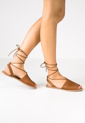 Brązowe skórzane sandały z otwartym przodem, z krzyżującymi się wokół kostek paskami na sznurówki i płaską podeszwą. Gładka faktura i prosty design.