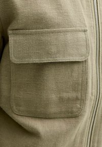 Nærbilde av en olivengrønn stofflomme med avrundede hjørner og klaff, sydd på en jakke ved siden av en sølvglidelås.