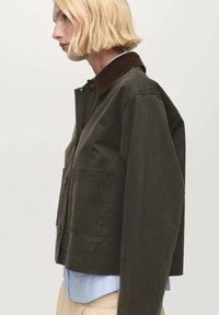 Veste olive verte coupée avec un col en velours côtelé, deux poches avant et une texture douce, portée sur une chemise bleu clair.
