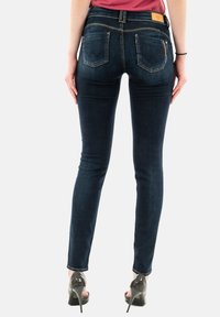 Jeans skinny azul oscuro con una cintura de altura media, bolsillos traseros con detalle de costura y cremalleras en las caderas, combinados con tacones altos transparentes.