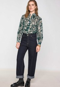 Blusa a fantasia floreale verde con colletto arricciato, abbinata a jeans dritti blu scuro con risvolti e cuciture a contrasto.