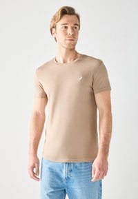 Camiseta de cuello redondo beige hecha de material suave, de mangas cortas, diseño ajustado, con un pequeño logo blanco en el lado izquierdo del pecho.
