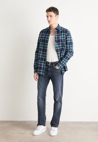 Camisa de franela para hombre en un patrón de cuadros en azul marino y verde, diseño abotonado, combinada con jeans oscuros y zapatillas blancas. Look sencillo y casual.
