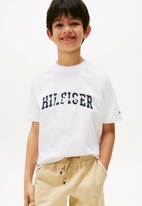 Hvid kortærmet t-shirt med navy "HILFIGER" logo, parret med beige shorts med snortræk i rød, hvid og blå nuancer.