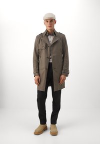 Trench coat color marrone chiaro con chiusura a bottoni e spalline, polo grigia, pantaloni neri e scarpe con tacco color tan. Sfondo semplice.