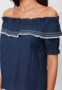 Top blu navy senza spalle con scollo arricciato, caratterizzato da dettagli ricamati bianchi e maniche corte arricciate. Realizzato in tessuto leggero.