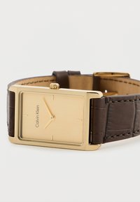 Χρυσό ορθογώνιο ρολόι με λείο, χρυσό καντράν, καφέ δερμάτινο λουρί με υφή από κροκόδειλο και μινιμαλιστικούς δείκτες ωρών.