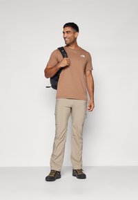 Camisa marrón de manga corta, pantalones cargo beige, mochila negra y zapatos de senderismo negros con cordones naranjas, de pie frente a un fondo gris.