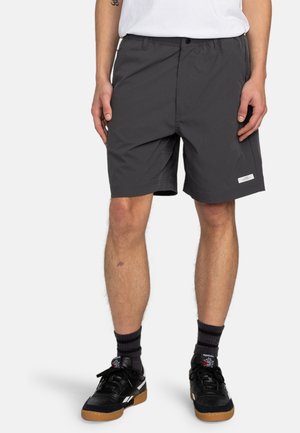 Gray shorts with an elastic waistband, side pockets, and a small logo tag on the hem. Worn with black sneakers and striped socks.