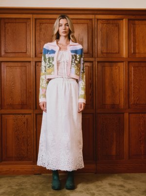 Ženska stoji ob lesenem stenovem ozadju, oblečena v belo čipkasto krilo, pastelno jopico s cvetličnim vzorcem in temno zelene gležnarje.