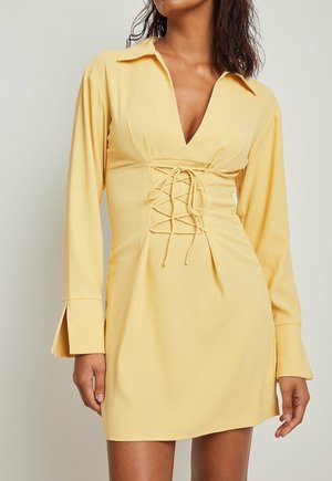 Gelbes Kleid mit V-Ausschnitt, langen Ärmeln und einer Schnürung vorne. Der Stoff hat eine glatte Textur, und die Ärmel sind leicht ausgestellt.