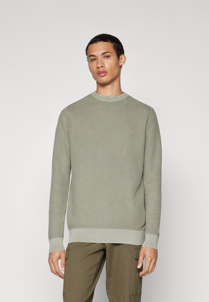 Helles grünes Strickpullover mit Strukturmuster, Rundhalsausschnitt und gerippten Bündchen. Dezentes Logodetail auf der Brust.