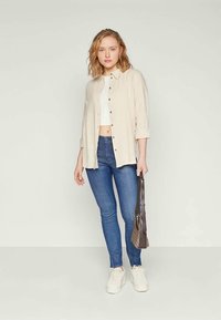 Beige knappskjorte, hvit crop top, blå skinny jeans og hvite sneakers; en liten mønstrete håndveske fullfører antrekket.