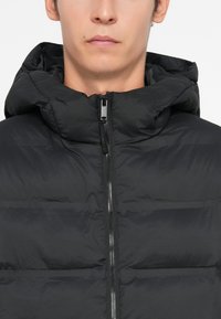 Mustaherkkävuori, jossa on huppu; edessä vetoketju ja vaakasuora tikattu tekstuuri. Valmistettu sileästä, kevyestä materiaalista.