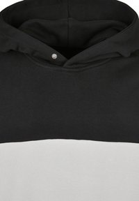 Sweat à capuche noir et blanc avec une partie supérieure noire muni d'une capuche et une partie inférieure blanche. Comprend un détail à bouton-pression argenté au niveau de l'encolure.