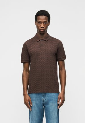 Junger Mann steht, trägt ein braunes Polohemd mit Muster und blaue Jeans, schaut nach vorne vor einem schlichten hellen Hintergrund.