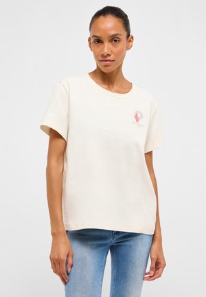 Frau trägt ein schlichtes, cremeweißes T-Shirt mit kurzen Ärmeln, verziert mit einer kleinen pinkfarbenen Kaktusgrafik und dem Schriftzug "Mustang" auf der Brust, kombiniert mit hellblauen Jeans.