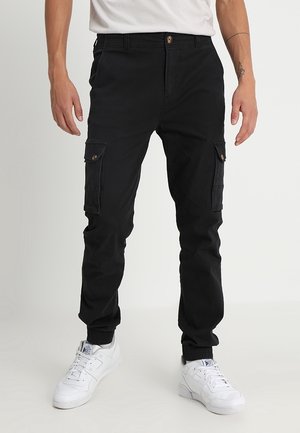 Homme portant un pantalon cargo jogger noir avec des poches latérales à boutons, un t-shirt blanc et des baskets blanches, debout devant un fond uni.