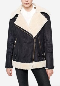 Kvinde iført en sort shearling-jakke med cremefarvet fleece-for, stor krave, lynlås foran og spænde-detaljer, parret med sorte bukser.