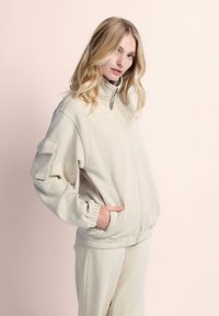 Beige zip-up jack met een hoge kraag, ribbelmanchetten en een patch pocket op de arm; zachte textuur en relaxed fit gecombineerd met bijpassende broek.