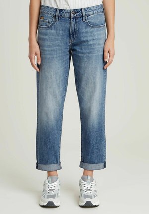 Persoon draagt rechte blauwe jeans met omgeslagen pijpen en witte sneakers, staand met ontspannen armen langs het lichaam tegen een effen achtergrond.
