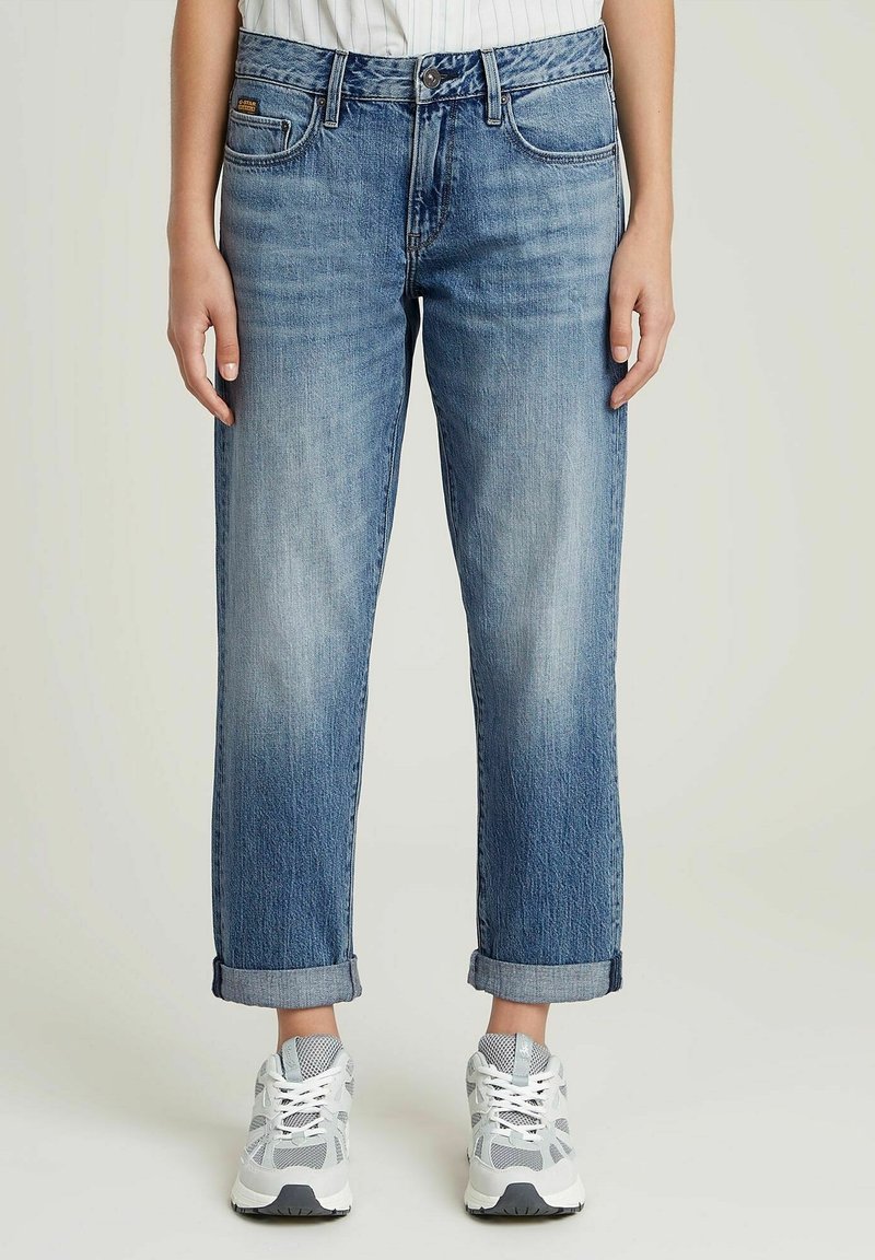 Personne portant un jean bleu droit avec revers et des baskets blanches, debout, les bras détendus le long du corps, sur un fond uni.