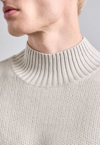 Pulover tricotat de culoare bej deschis, cu guler înalt cu nervuri și un corp texturat, evidențiind cusături fine și un material moale, respirabil.