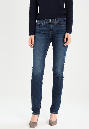 Vrouw die donkerblauwe slim-fit jeans, een marineblauwe top met lange mouwen en grijze suède puntige hakken draagt, staand tegen een effen witte achtergrond.