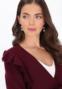 Burgundowy kardigan z falbaniastymi akcentami, żebrowaną strukturą i głębokim dekoltem w kształcie V. Połączony z białą bluzką i perłowymi kolczykami z wiszącymi elementami.