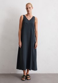Robe maxi sans manches de couleur foncée avec un décolleté en V, silhouette en A fluide, fabriquée dans un tissu lisse. Assortie à des sandales glissantes noires avec accessoires en or.