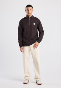 Dark brown zip-up sweatshirt with a high collar, featuring a logo on the chest, paired with cream-colored pants and light sneakers.