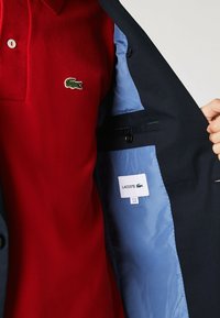 Röd polo tröja i texturerat tyg med krage, med en grön krokodillogotyp. Inom en marinblå kavaj med ljusblå foder och etikett.