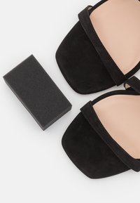 Paire de sandales ouvertes en daim noir avec des brides à la cheville à côté d'une éponge à chaussures noire rectangulaire sur fond blanc.
