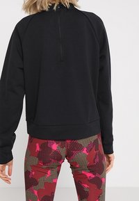 Czarny sweter z wysokim kołnierzem i zamkiem z tyłu, w połączeniu z wzorzystymi legginsami w odcieniach czerwieni, różu i brązu z siatkowym wzorem.