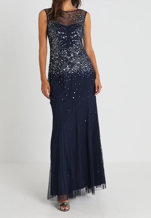 Robe de soirée sans manches bleu marine avec haut transparent au niveau de la poitrine, ornée de sequins argentés, portée avec des talons ouverts argentés sur fond blanc.