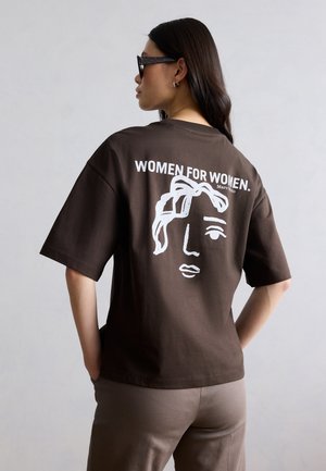 Γυναίκα που φορά καφέ πουκάμισο με λευκό αφηρημένο πρόσωπο και κείμενο «ΓΥΝΑΙΚΕΣ ΓΙΑ ΓΥΝΑΙΚΕΣ.» στην πλάτη, στέκεται μπροστά σε απλό ανοιχτό γκρι φόντο.