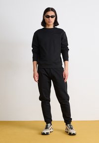 Svart sweatshirt med långa ärmar och ribbade muddar, tillsammans med matchande joggers. Svarta solglasögon och krämfärgade sneakers med svarta accentfärger.