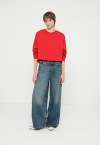 Czerwony krótki sweter z długimi rękawami w połączeniu z jasnoniebieskimi spodniami typu wide-leg. Model nosi białe trampki. Jednolity kolor, casualowy fason.
