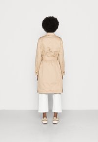 Trench coat beige con vita regolabile, maniche lunghe e polsini con bottoni, abbinato a pantaloni bianchi larghi e sneakers chiare.