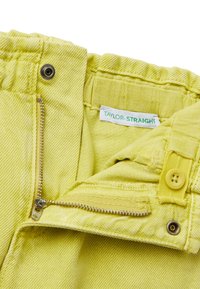 Žluté džíny rovného střihu s předním zipem, knoflíkovým zapínáním a označením značky. Texturovaná tkanina s viditelným švem a detaily kovových komponentů.