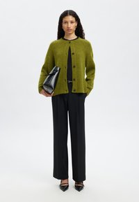 Žalias megztas kardiganas su sagomis, derinamas su juodomis kelnėmis ir juodais aukštakulniais su smailiais galiukais. Laikoma juoda struktūruota rankinė.