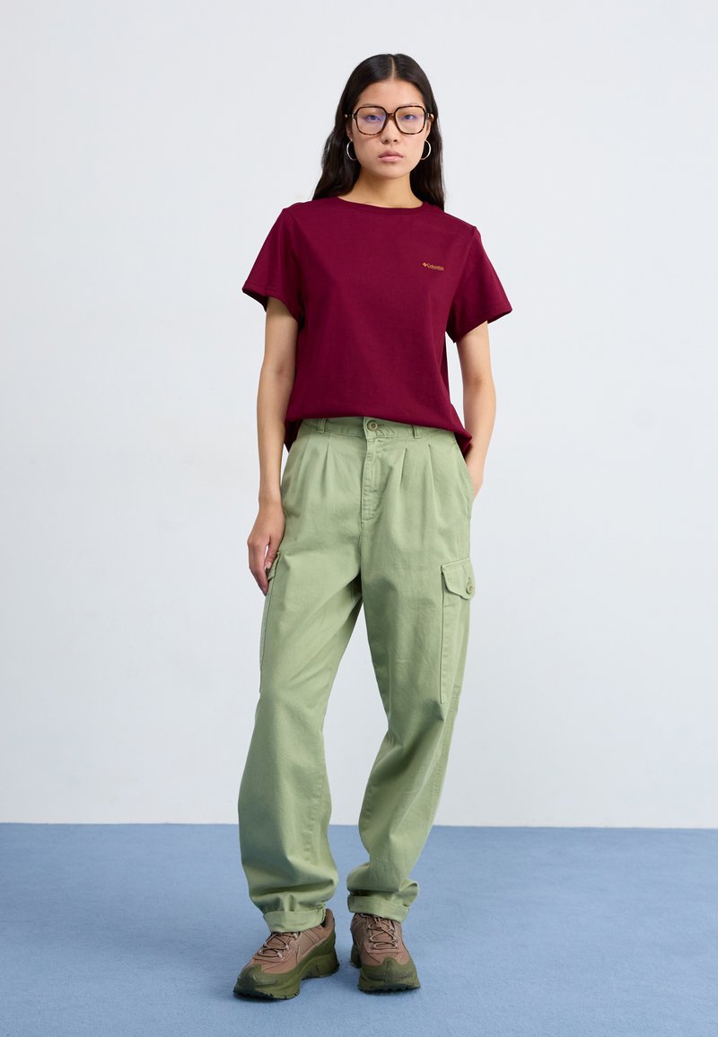 Bordowa bawełniana koszulka z krótkim rękawem, zestawiona z jas zielonymi spodniami cargo z dużymi kieszeniami i luźnym krojem. Brązowe buty trekkingowe.