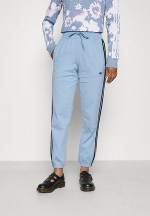 Person wearing light blue Adidas track pants with black side stripes, floral long-sleeve shirt, and black leather shoes with buckles.