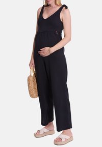 Schwarzer, ärmelloser Umstandsoverall mit Bindegurten, aus weichem Textilmaterial und in lockerer Passform. Eine gewebte Tasche und flache Sandalen runden das Outfit ab.