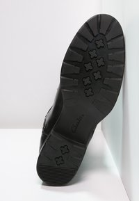 Semelle de chaussure en cuir noir avec un motif de crampons profonds comprenant des formes croisées et des rainures rectangulaires épaisses, présentée sur un fond blanc.