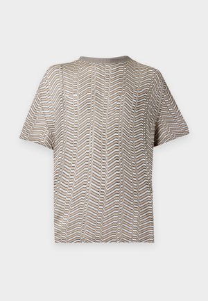 Mann steht, trägt ein kurzärmeliges Hemd mit Chevron-Muster, hellbraune Hose und dunkelbraune Lederschuhe, Hände in den Taschen.