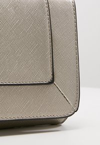 Beżowa torebka o fakturowanej powierzchni z geometrycznym wzorem, ozdobiona czarną lamówką i przeszyciami. Projekt cechuje się uporządkowanym kształtem i ostrymi kątami.