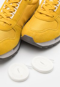 Sneaker gialli realizzati in suede e mesh con una finitura testurizzata, dotati di lacci bianchi e suole in gomma grigia, accompagnati da lacci bianchi arrotolati.