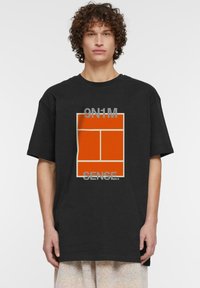 Juoda per didelė marškiniai su oranžiniu grafiniu dizainu, kuriame yra tekstas "9N1M" ir "SENSE." Įprasta medvilnės medžiaga su minkštos tekstūros.